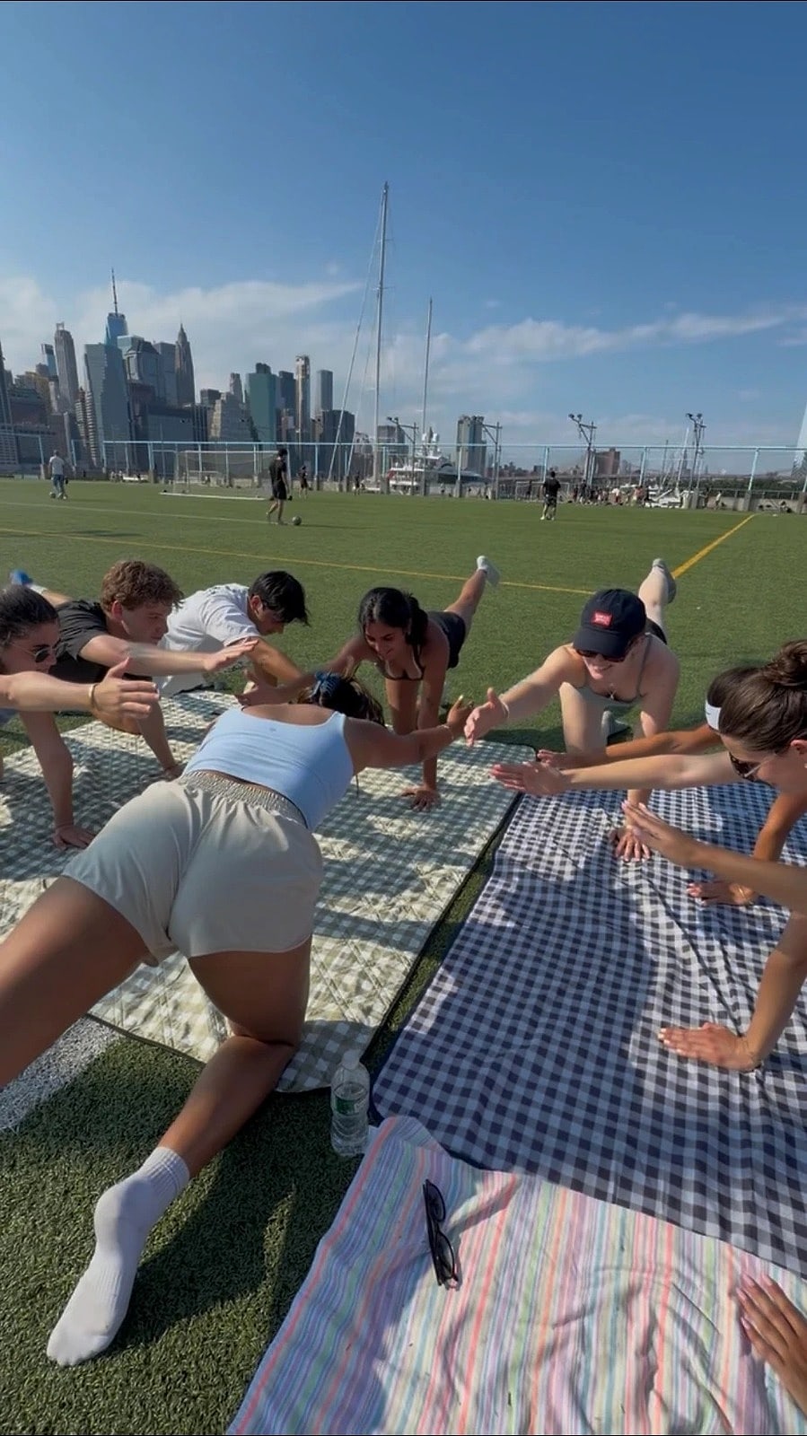 An outdoor class on picnic blankets on a waterfront lawn, with the Manhattan skyline behind