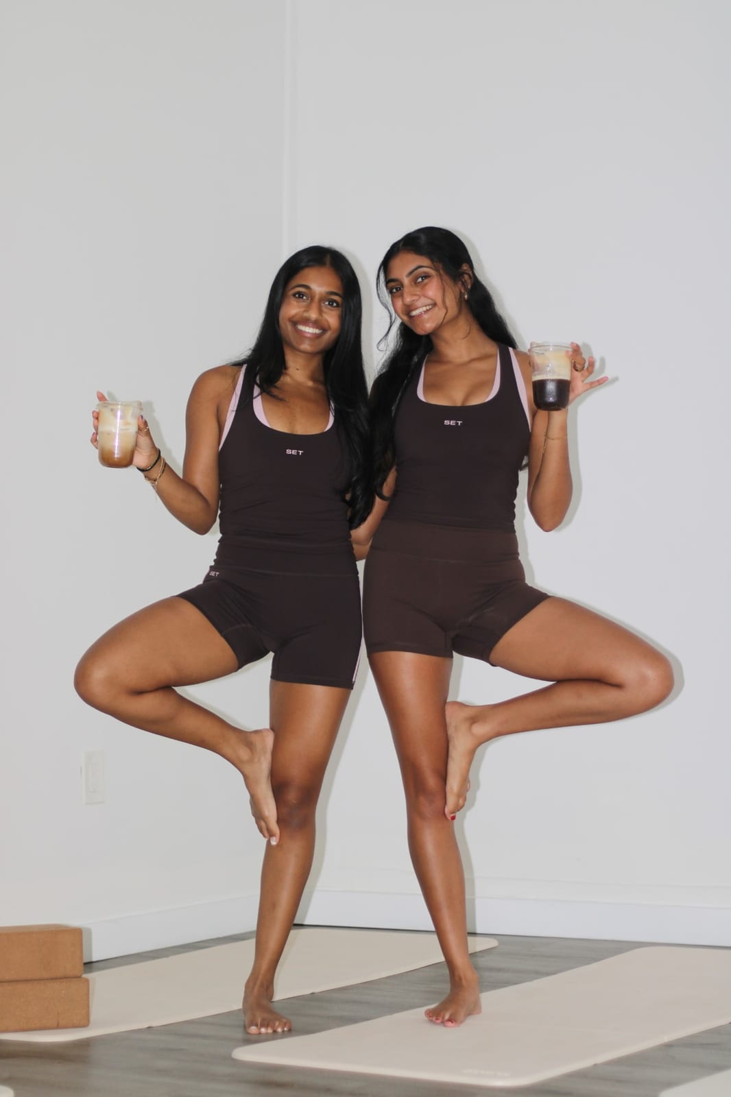 Two women in matching outfits doing tree pose, each holding an iced coffee