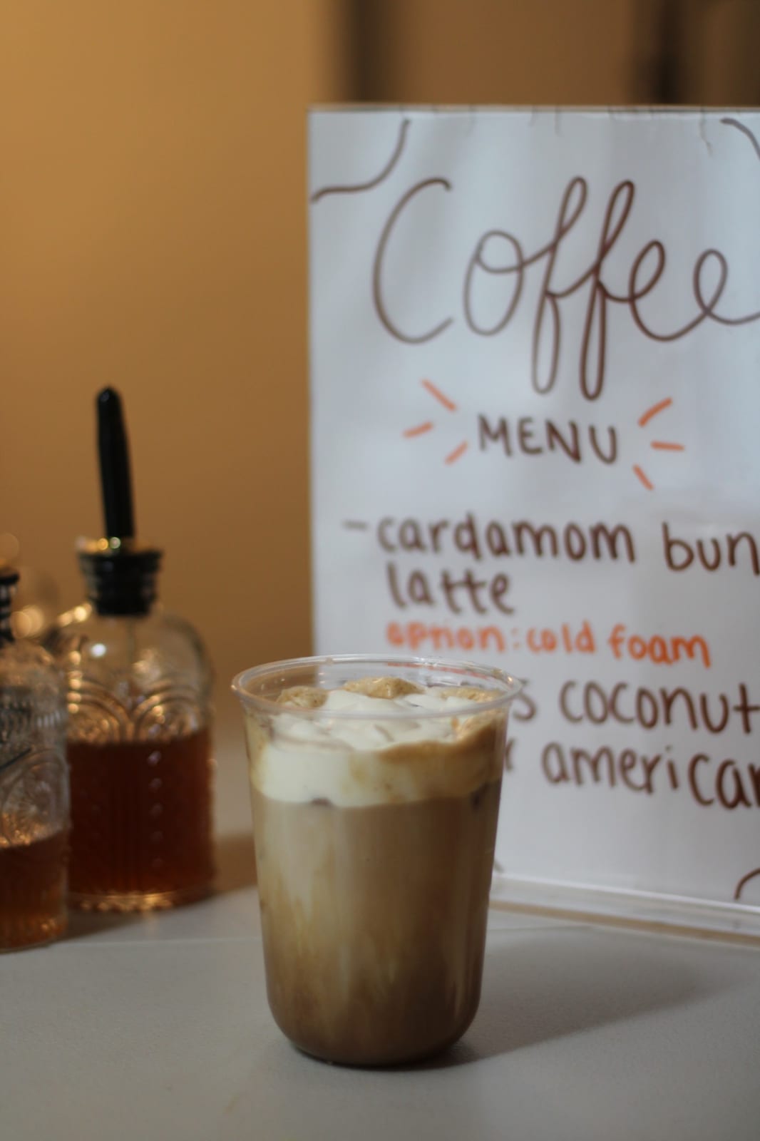 An iced cardamom bun latte with cold foam in front of the handwritten menu
