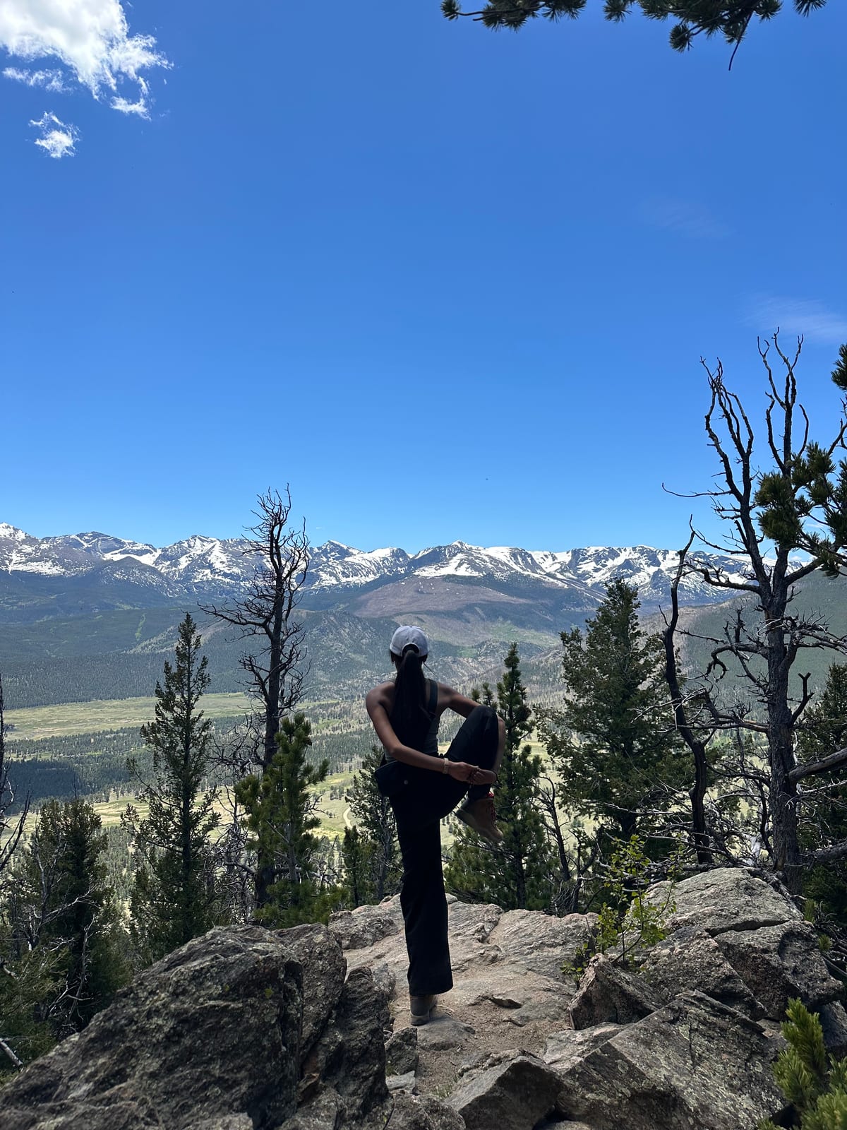 Saniya in tree pose on a rocky outcrop above a forested valley and snowy peaks