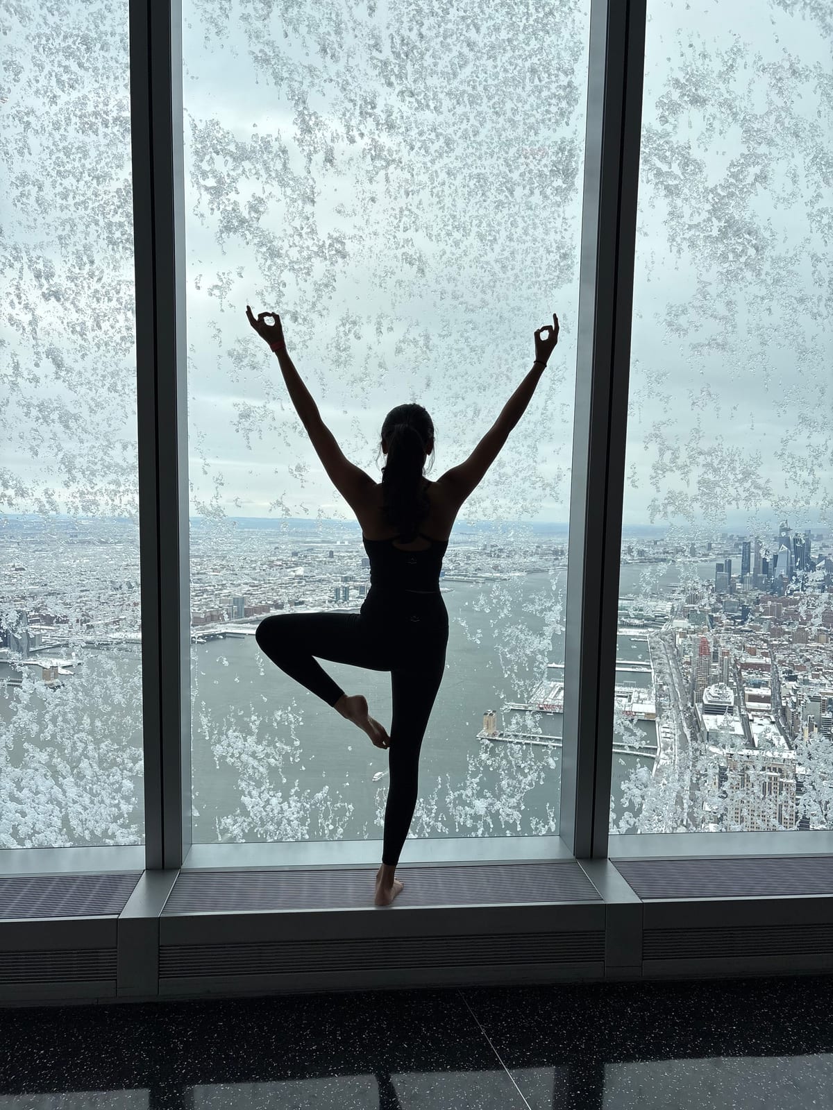 Saniya balancing with arms raised at a floor-to-ceiling window high above the city