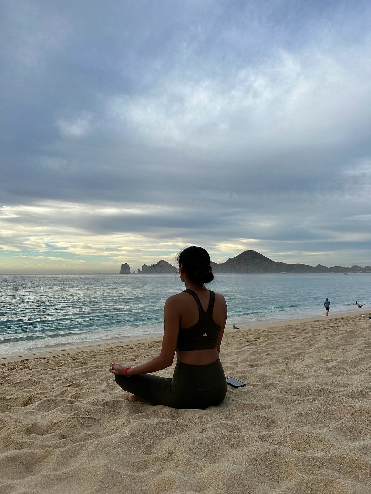 Saniya meditating cross-legged on the sand, facing the ocean and rocky headland at dawn