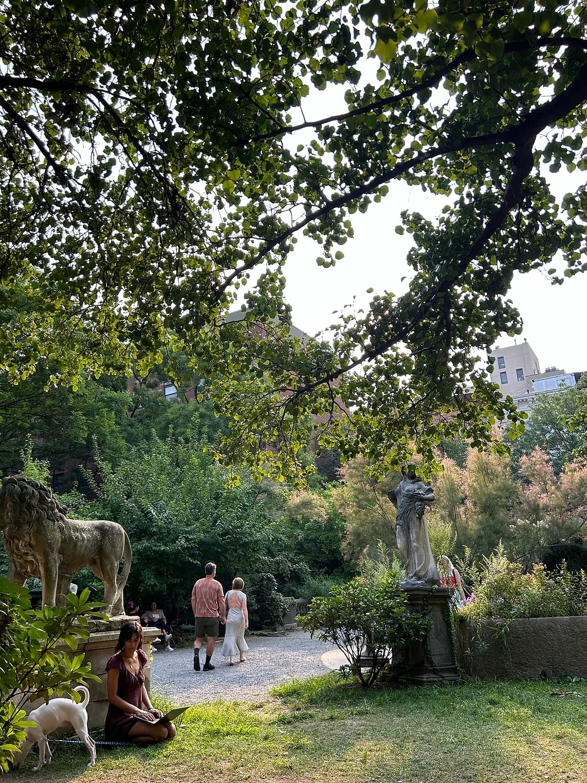 A leafy garden path lined with stone statues, with people sitting on the grass