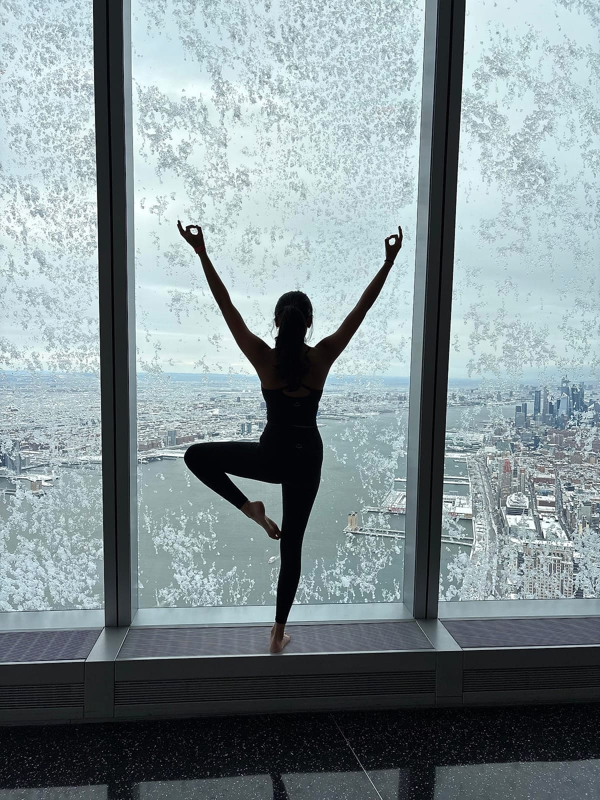 Saniya balancing with arms raised at a floor-to-ceiling window high above the city